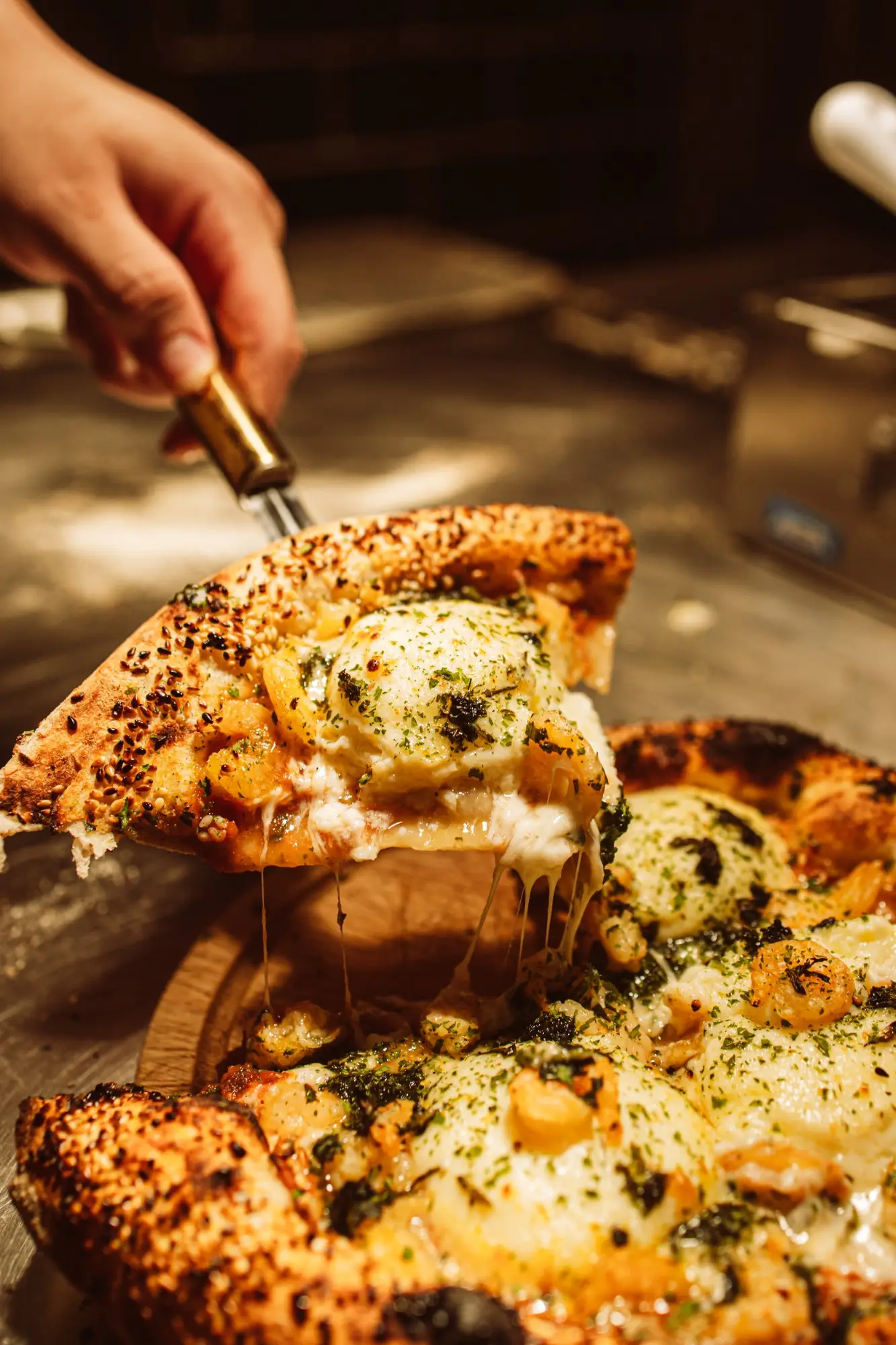 Fatia de pizza de camarão sendo levantada, com o queijo esticando e a borda coberta de gergelim tostado