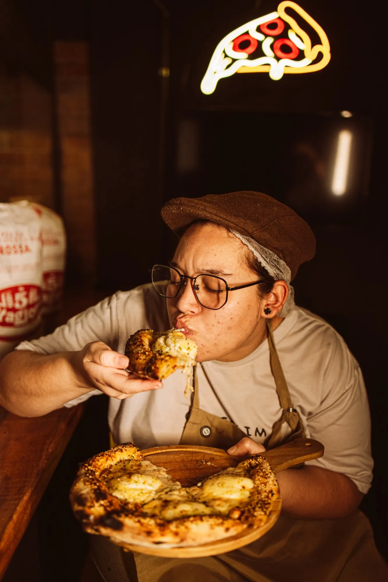 Pizzaiola da Gergelim provando uma fatia, de olhos fechados, sob o neon em forma de pizza