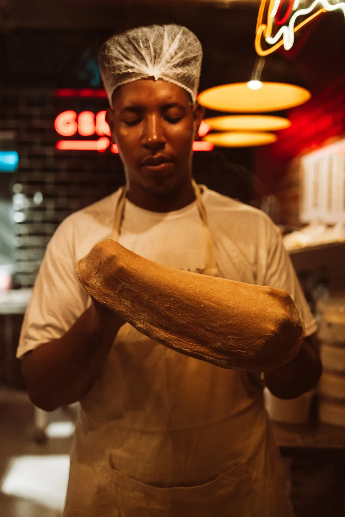 Pizzaiolo da Gergelim segurando um bloco de massa na cozinha, com o neon da marca ao fundo