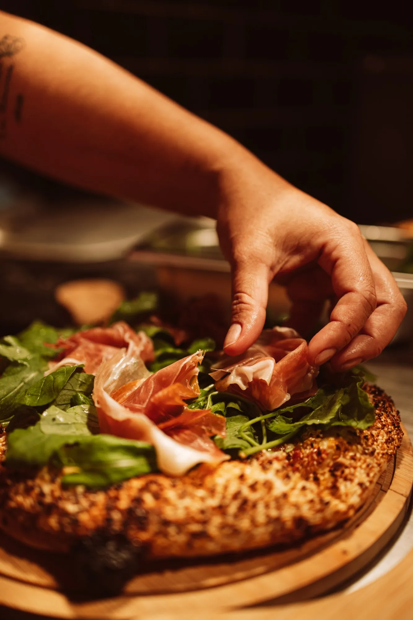 Mão finalizando uma pizza com fatias de presunto parma sobre rúcula fresca