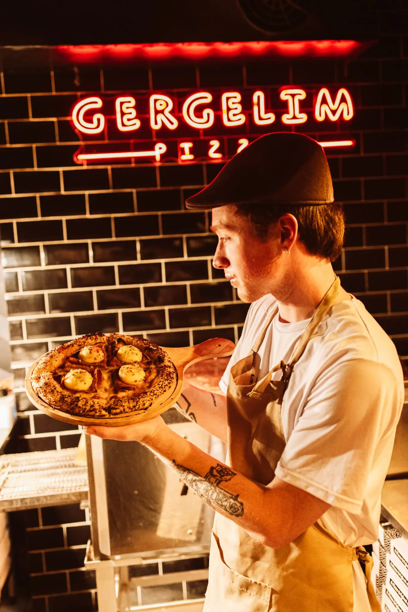 Pizzaiolo segurando uma pizza pronta diante do letreiro em neon da Gergelim