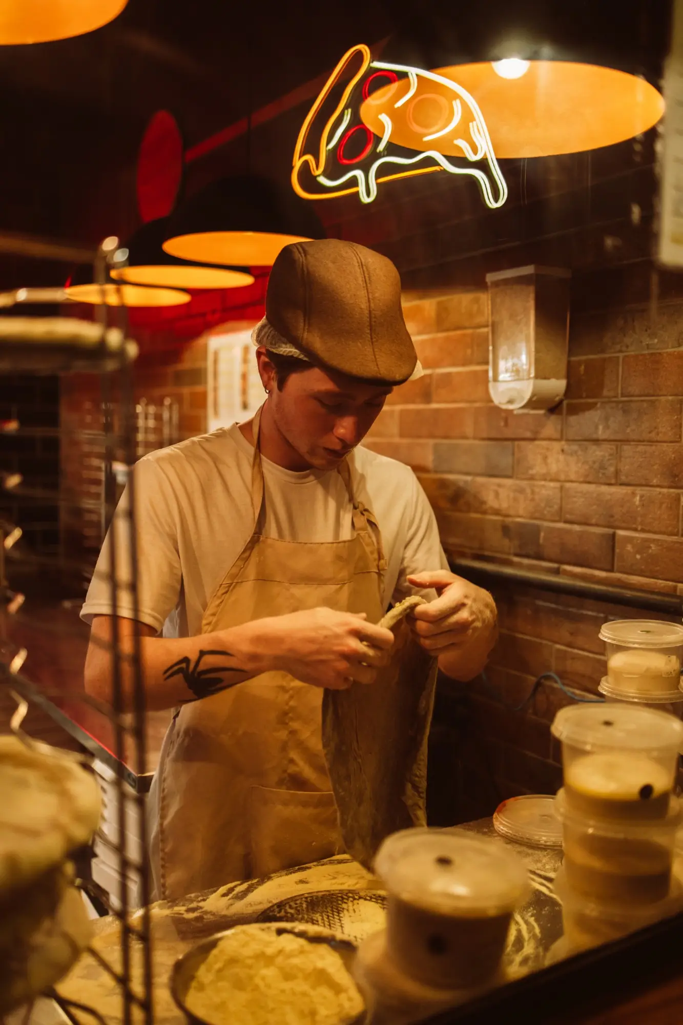 Pizzaiolo abrindo a massa à mão ao lado dos potes de fermentação, sob o neon em forma de pizza